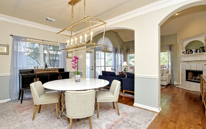 Open-plan dining and living room with round white table, cream chairs, brass pendant light, and arched doorways to adjacent rooms.
