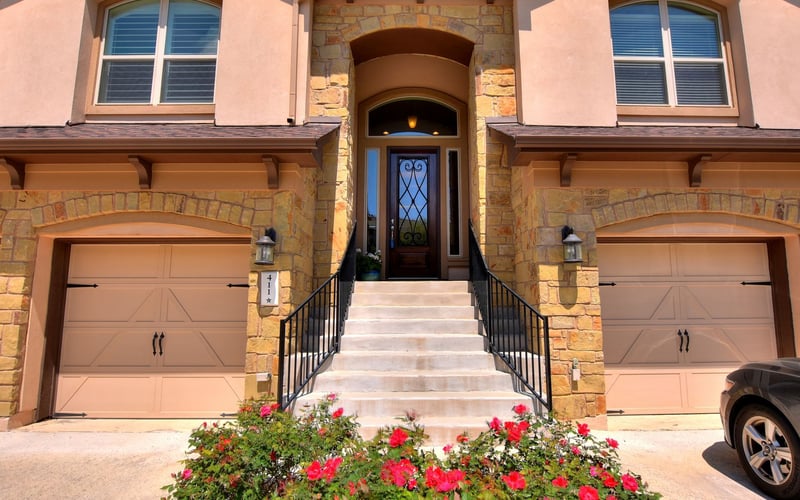 Two-story stone and stucco house with arched entryway, dual garage doors, and pink flowering shrubs in front
