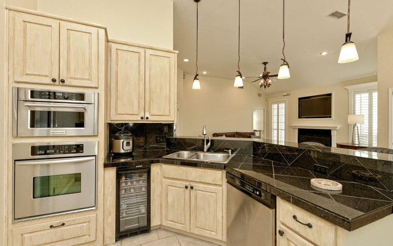 Open kitchen with light wood cabinets, dark granite countertops, stainless steel appliances, and pendant lights overlooking living room with fireplace.