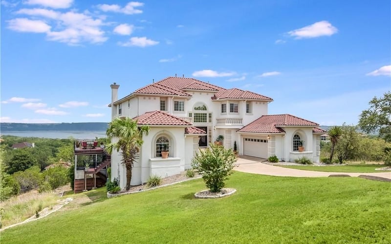 Two-story white villa with terracotta tile roof, manicured lawn, and lake view in background